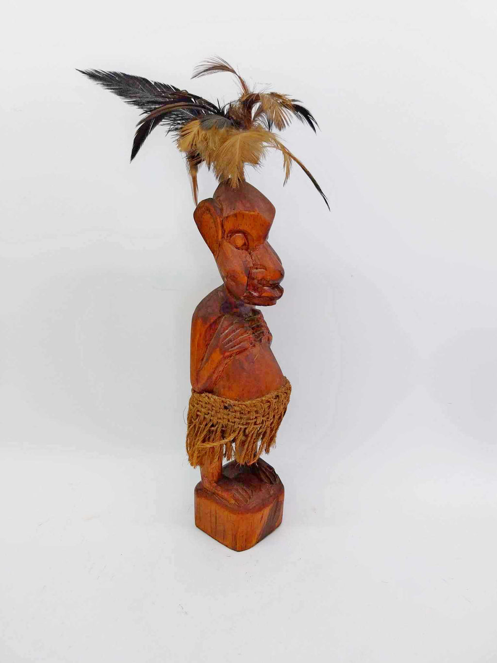 Vintage amber hardwood tribal carved figure with feather headdress and raffia skirt on white background