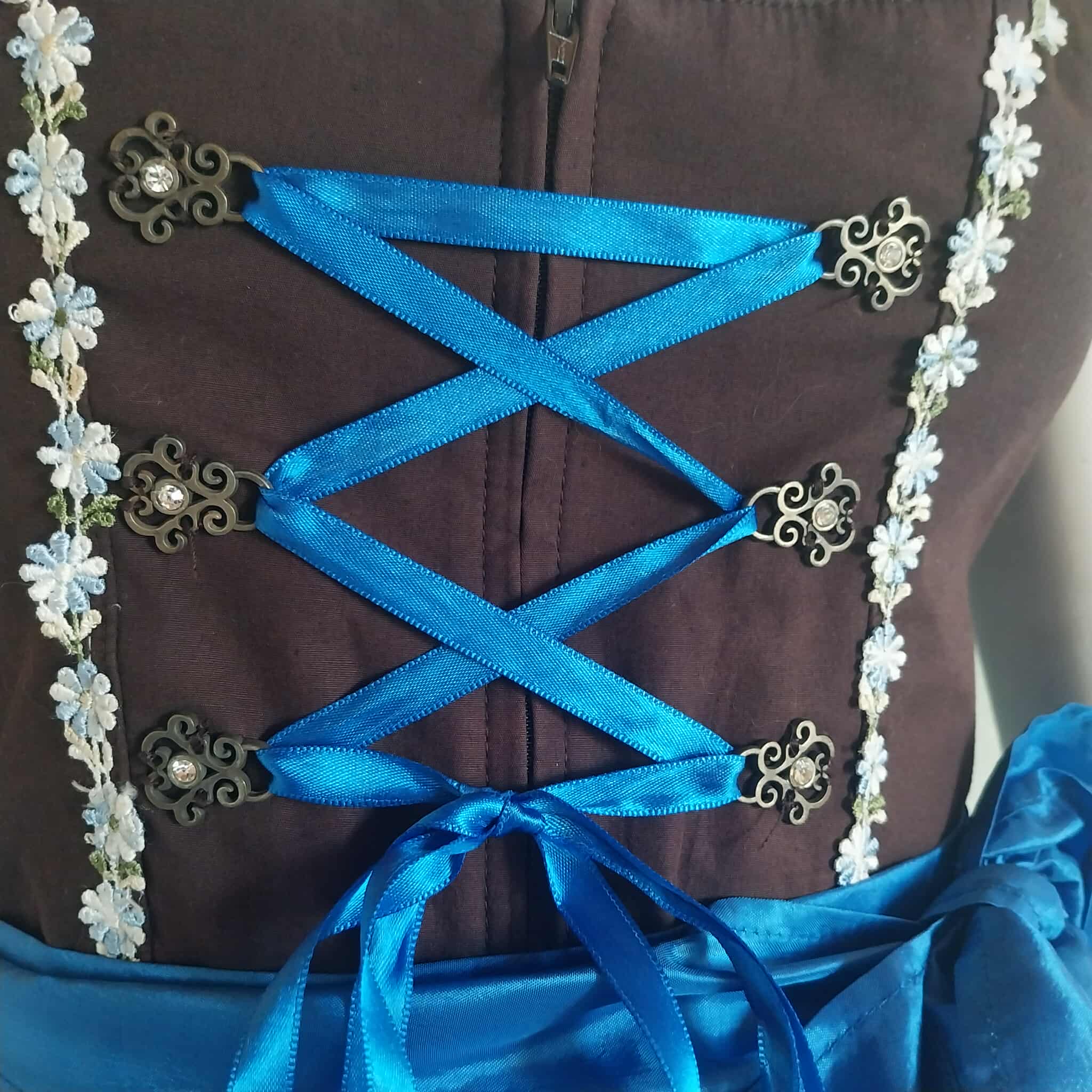 Close-up of Stockerpoint dirndl bodice detail — blue satin cross-lace ribbon with bronze rhinestone clasps and floral trim