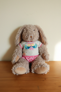 Moulin Roty fluffy plush rabbit with owl t-shirt and pink heart shorts, sitting on warm wooden surface